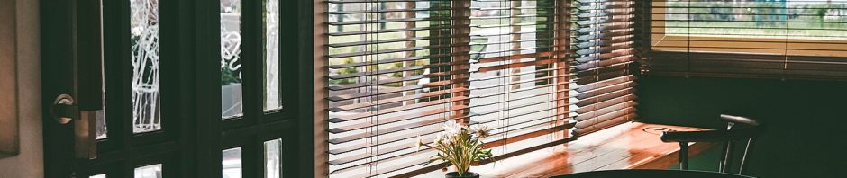 Wooden venetian blinds on a café window with a counter, stools, and a small potted plant in warm light.